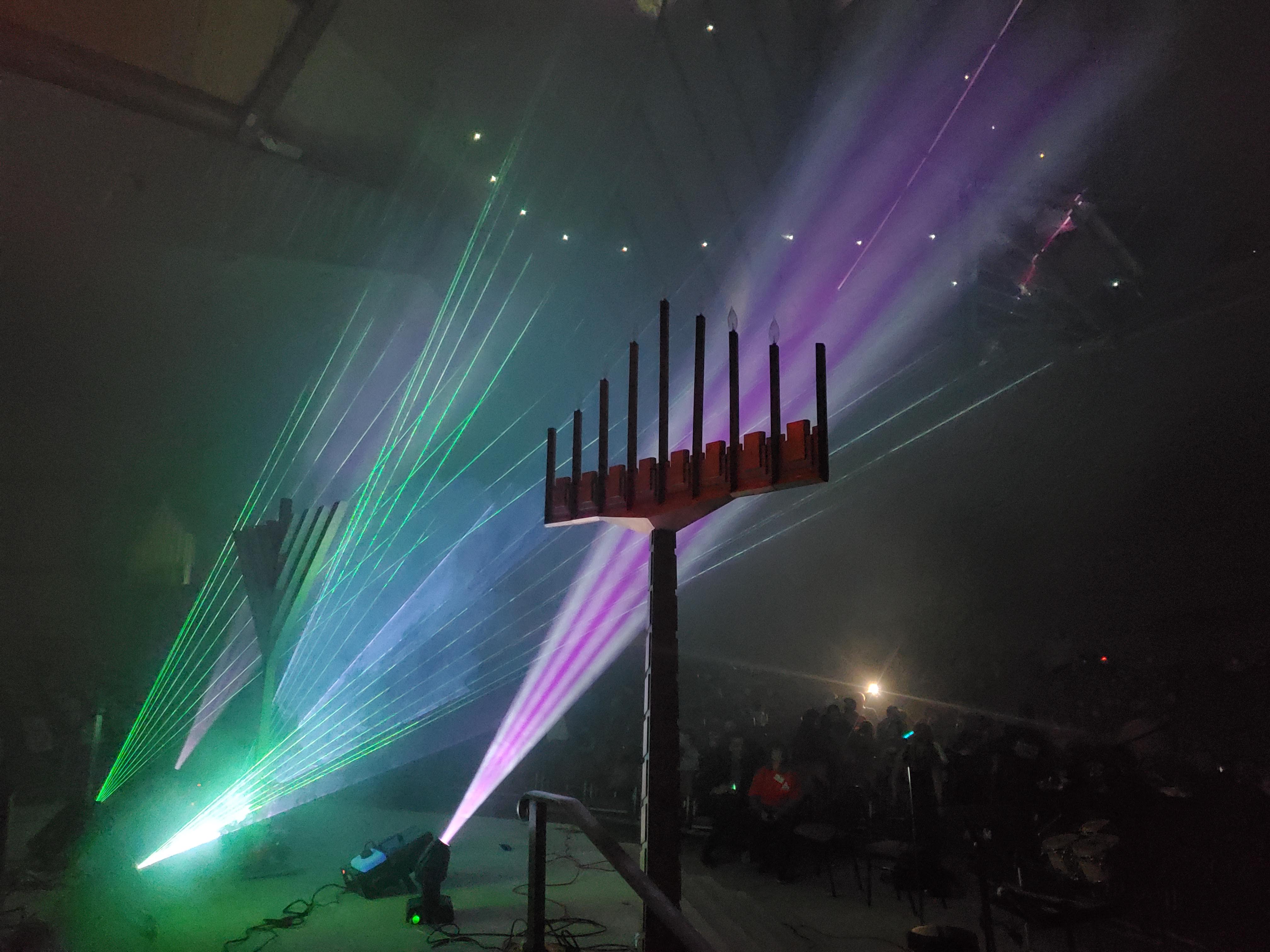 A tall menorah structure standing inside multicolored laser beams and haze