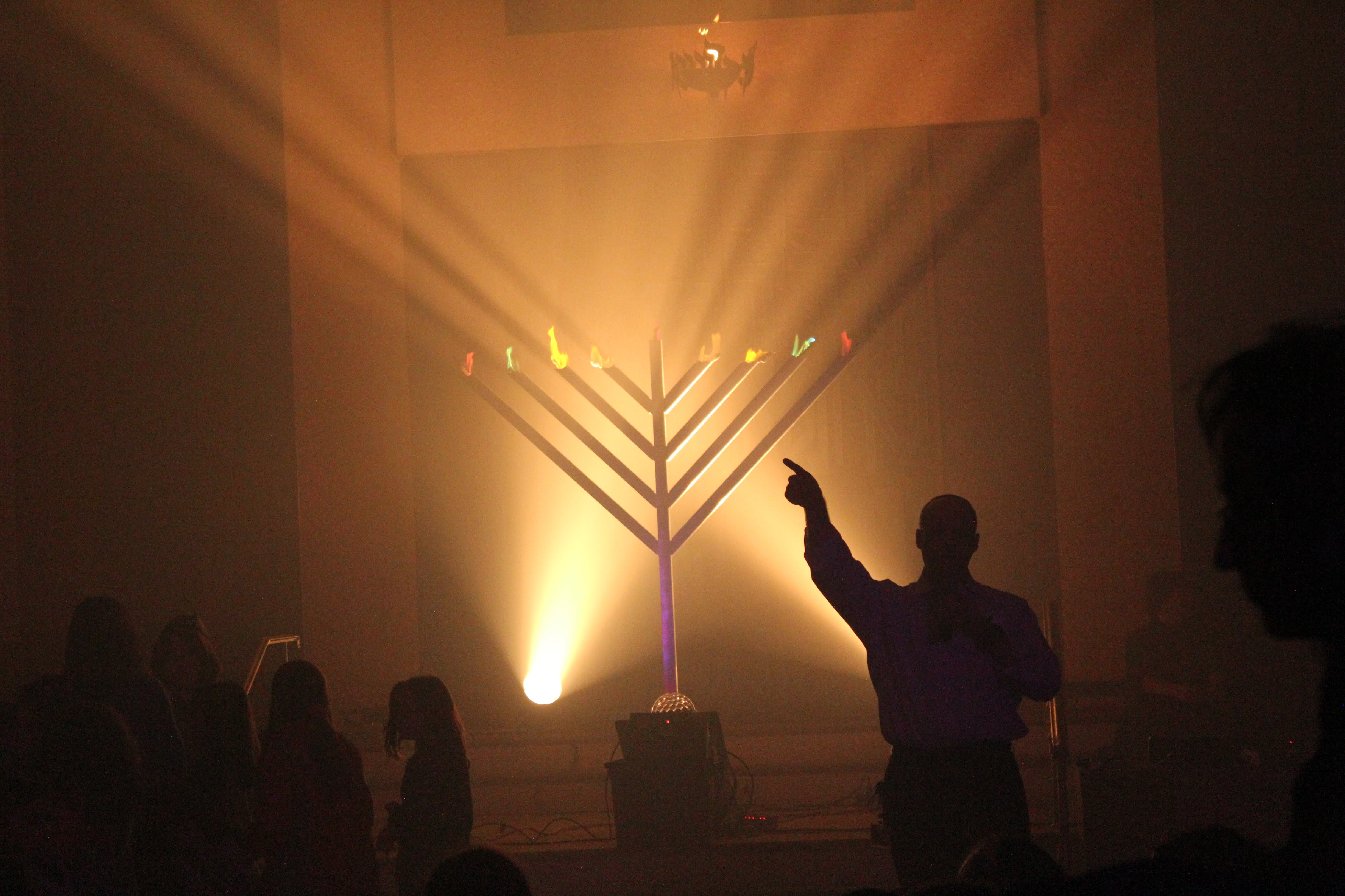 A giant glowing menorah with a person reaching up toward the many-colored flames