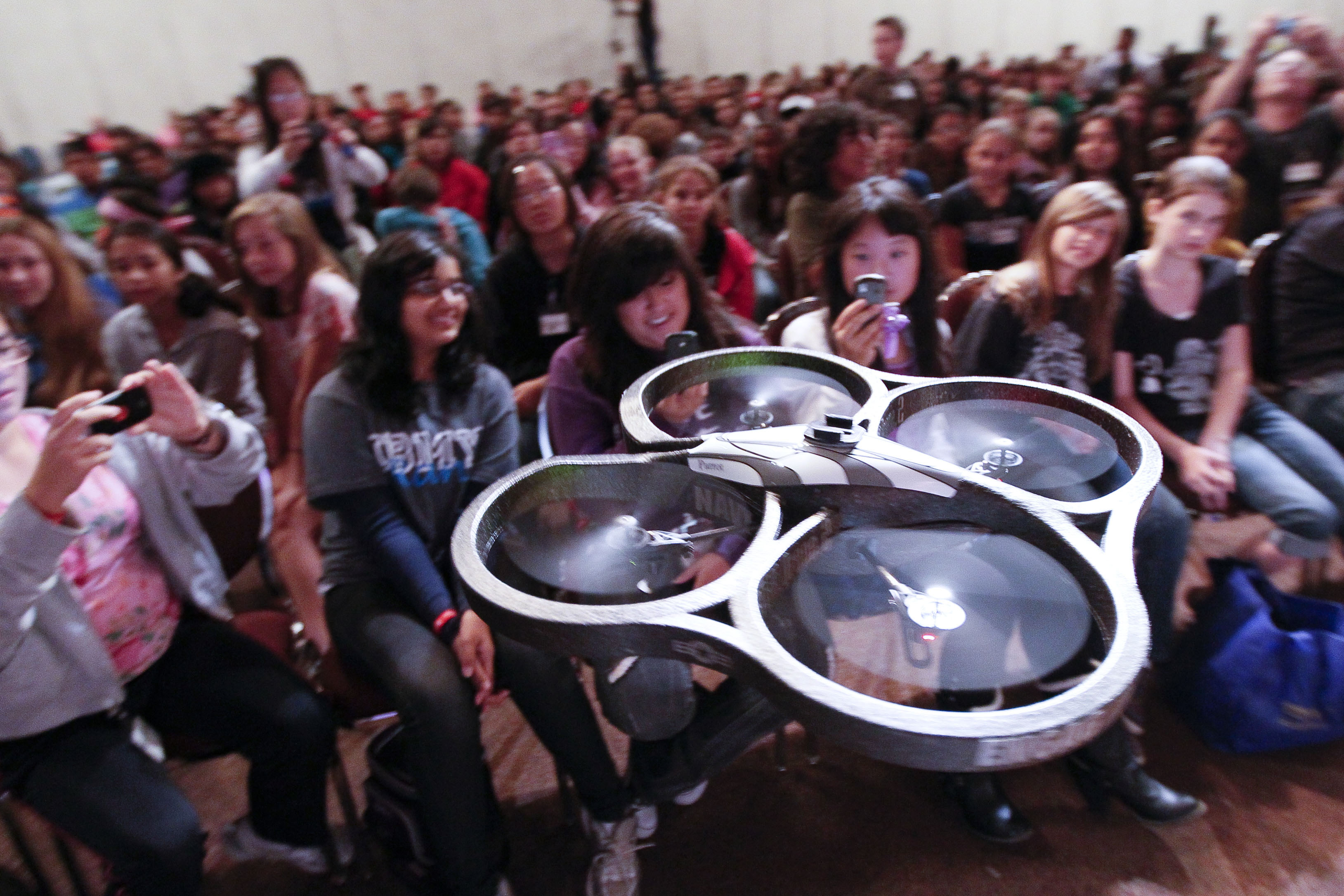 A Parrot AR drone hovers inches from a row of delighted teen girls in a packed school auditorium — hundreds of students visible behind them