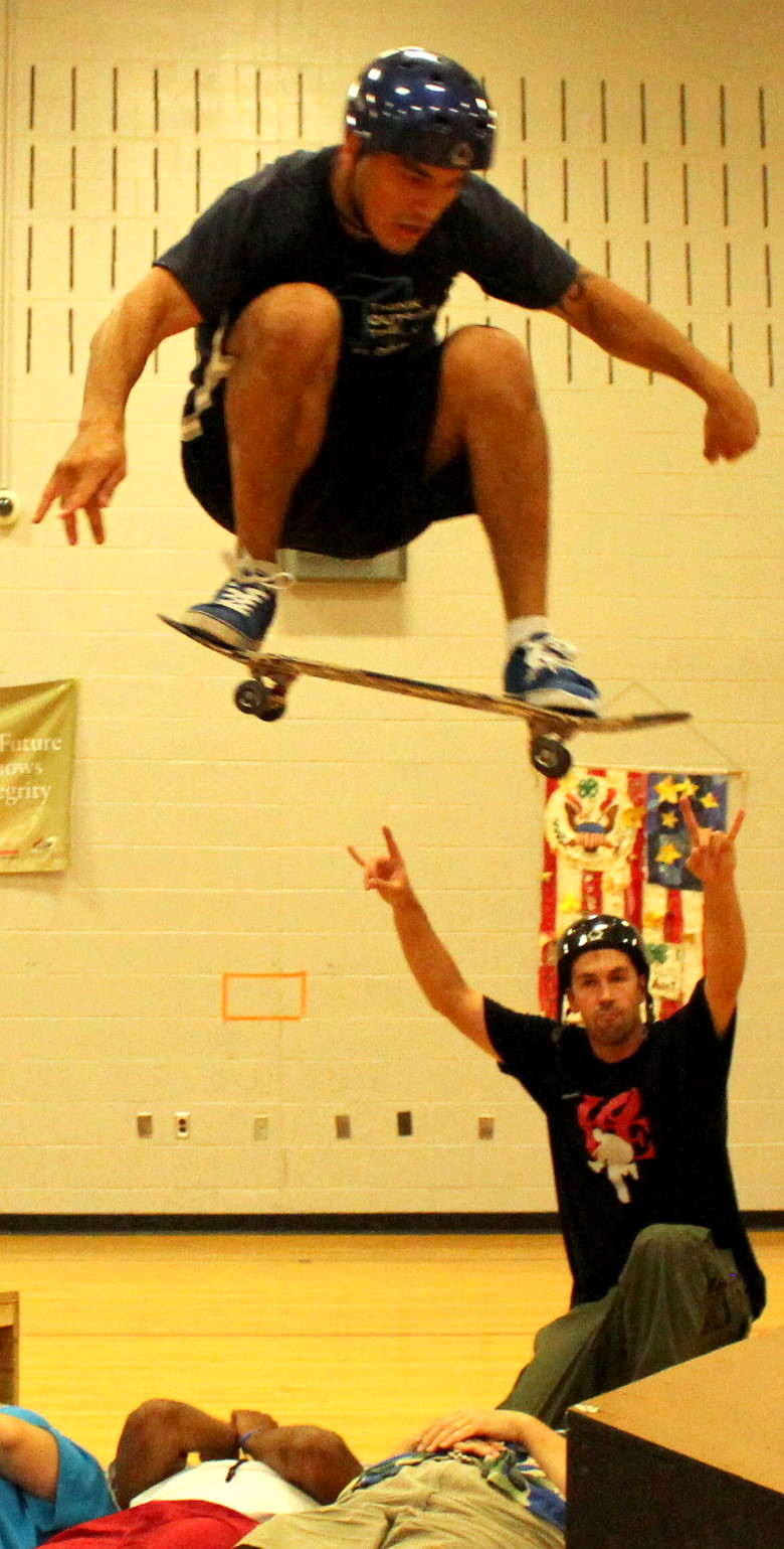 A skateboarder mid-air in a full board-grab trick above a school gymnasium floor, a second performer below with arms raised in celebration