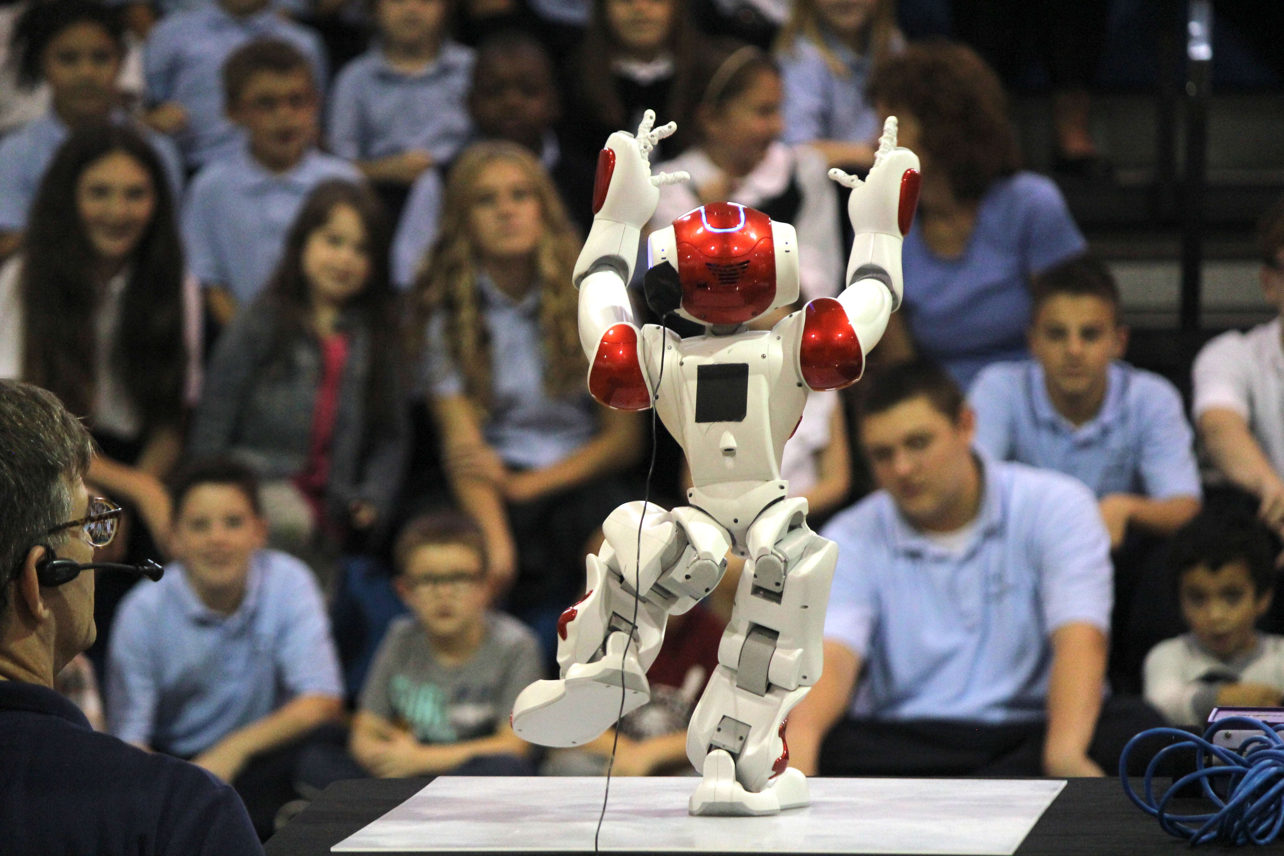 A NAO humanoid robot stands mid-dance with arms raised in a school gymnasium, a packed auditorium of students behind it
